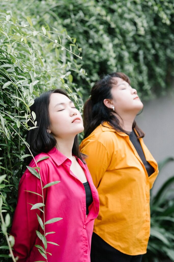 Two women peacefully standing outdoors with eyes closed, surrounded by lush green leaves.