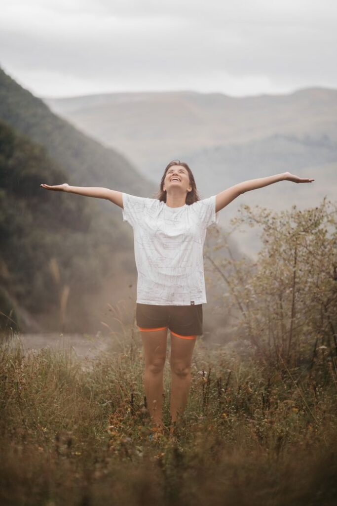 A woman stands joyfully with arms open in a lush mountain landscape.